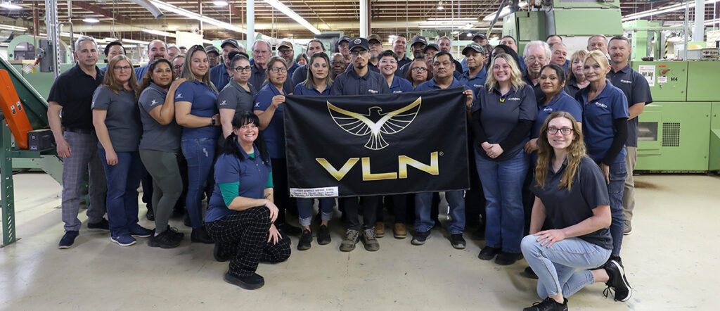 A group of workers in a factory pose together holding a black VLN banner with a gold bird logo.